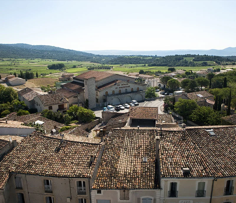 Vue aérienne (800x690) de la cave coopérative 'Les Coteaux de Montpeyroux' nichée parmi les toits de tuiles du village, avec la plaine viticole et les collines en arrière-plan. Montre le bâtiment de production dans son cadre.