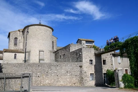 Les batiments du domaine : ancien prieure/eglise romane en pierre et murs anciens, visiteurs sur une terrasse a droite, ciel bleu. Architecture historique du domaine.