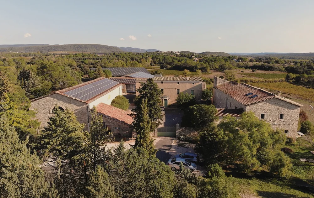 Vue aerienne d'ensemble du domaine: mas en pierre, cour interieure, toits, vignes et garrigue alentour avec collines a l'horizon. Excellente vue d'ensemble du chateau.