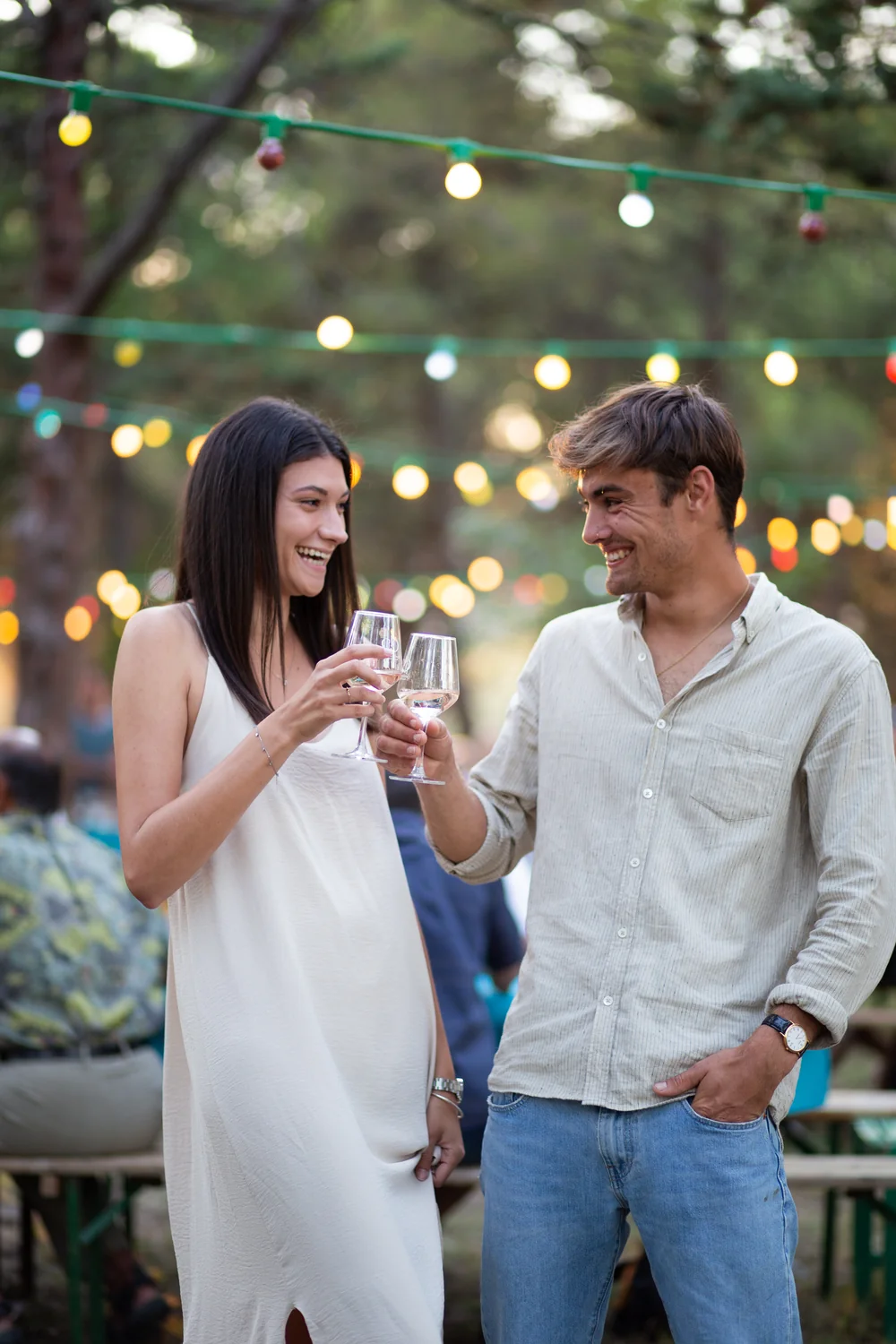 Couple souriant qui trinque avec des verres de blanc lors d'une soiree en exterieur, guirlandes lumineuses dans les pins. Scene d'evenement (Sunset Pic) chaleureuse et humaine.
