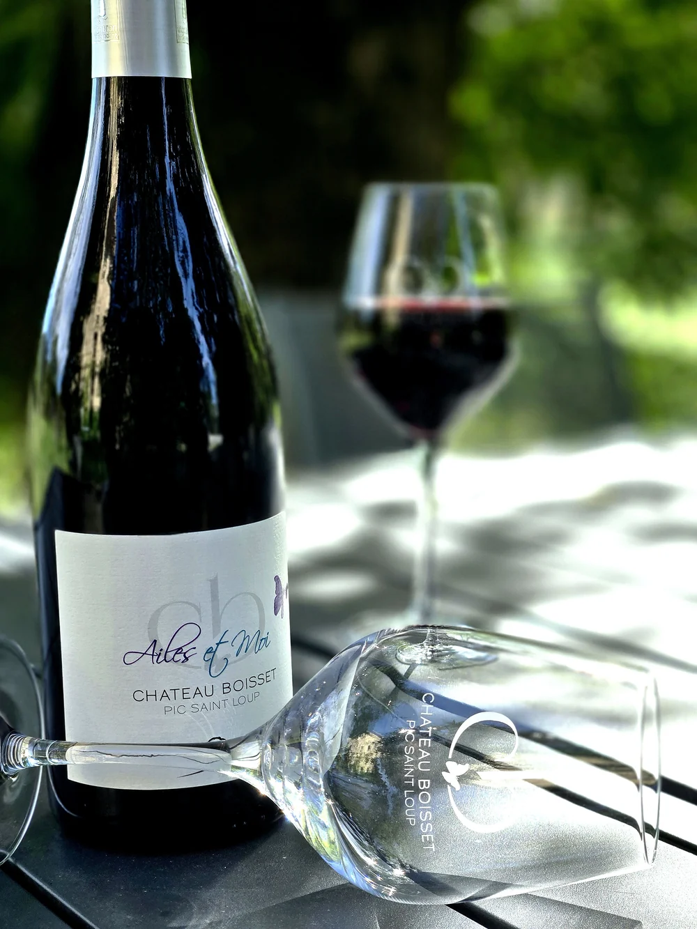 Bouteille cuvee 'Ailes et Moi' Pic Saint-Loup posee sur table avec verre de rouge en arriere-plan flou, jardin. Packshot lifestyle soigne.