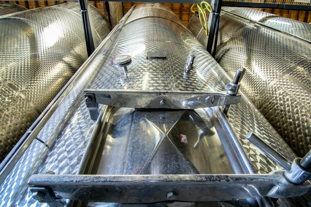 Cuves de vinification inox alignees dans le chai, vue plongeante sur les couvercles et la passerelle. Belle image technique du chai, lumineuse.