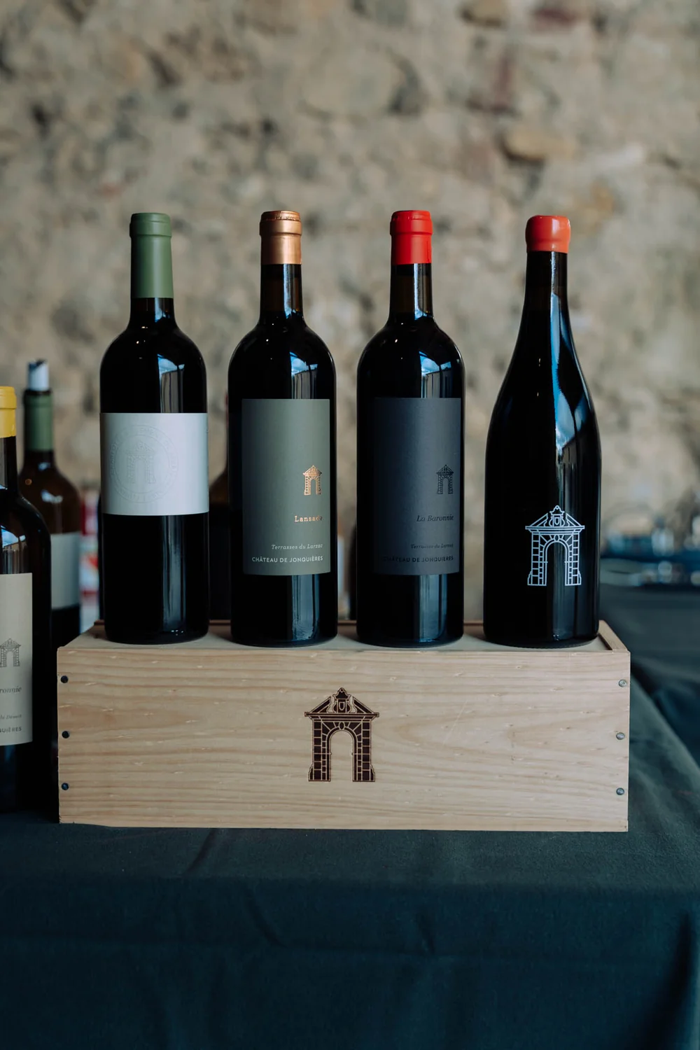 Packshot lifestyle professionnel (Melody Pallas) : quatre bouteilles de la gamme (Lansade, La Baronnie...) alignees sur une caisse en bois gravee du monogramme portail, devant un mur de pierre. Mise en scene soignee et premium.