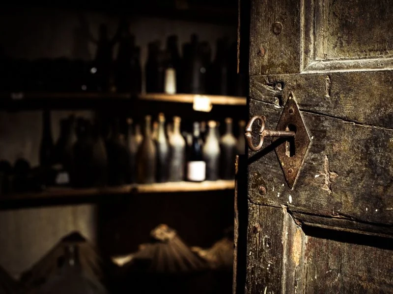 Photo d'ambiance du chai : vieille porte en bois entrouverte avec une cle ancienne dans la serrure en fer forge, vieilles bouteilles poussiereuses sur les etageres en arriere-plan flou. Atmosphere patrimoniale et intimiste, tres reussie.