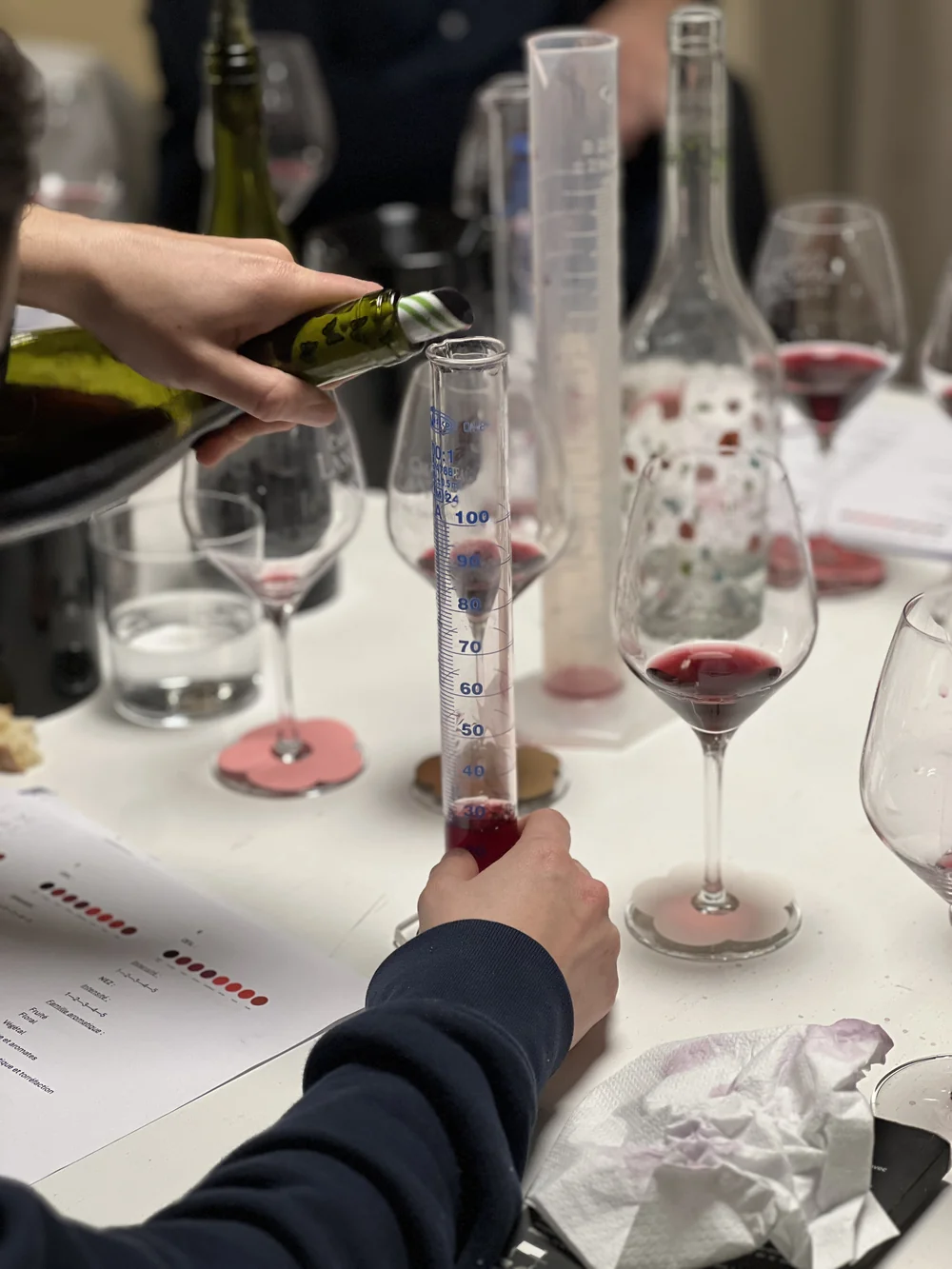 Gros plan sur une session d'assemblage/laboratoire : des mains versent du vin rouge dans une eprouvette graduee, verres de degustation et fiches autour. Montre le travail technique du chai.