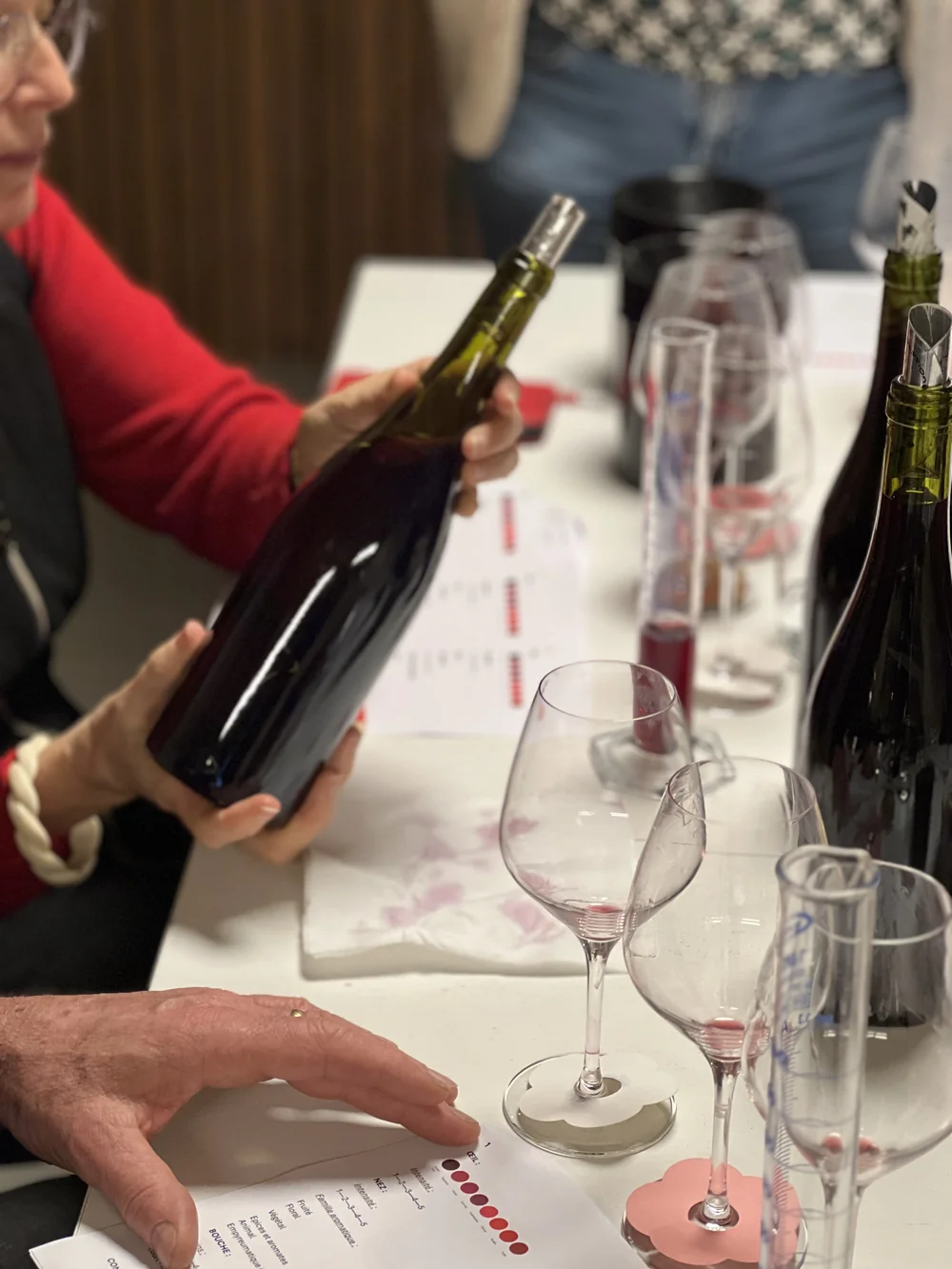 Atelier de degustation/assemblage : une personne en pull rouge tient un magnum, verres de degustation et fiches sur la table. Scene humaine d'atelier dans l'esprit du domaine.
