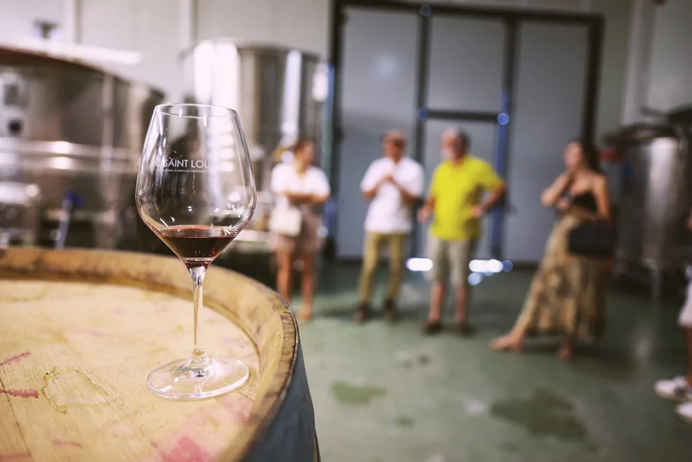 Verre de vin rouge (verre logote 'Pic Saint-Loup') pose sur une barrique au premier plan net, avec un groupe de visiteurs en degustation flou en arriere-plan dans le chai, devant les cuves inox. Belle profondeur de champ, ambiance oenotouristique conviviale. Cave + presence humaine. 1000x667.