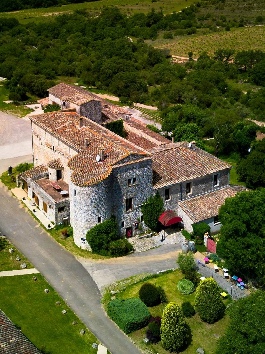 Vue aerienne rapprochee de tout le mas fortifie : tour ronde, toitures en tuiles, cour, allee bordee de parasols colores, vignes a l'arriere. Magnifique plan d'ensemble du domaine.