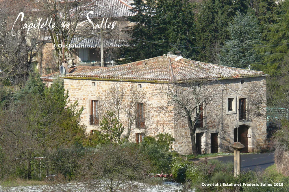Vue d'ensemble du mas familial en pierre du domaine, surnomme "L'Oustal" : belle batisse languedocienne en pierre seche avec toiture en tuiles, balcon en fer forge, entouree d'arbres et de garrigue. Photo prise en hiver/fin d'hiver. Filigrane calligraphie "Capitelle des Salles" + mention "L'Oustal"