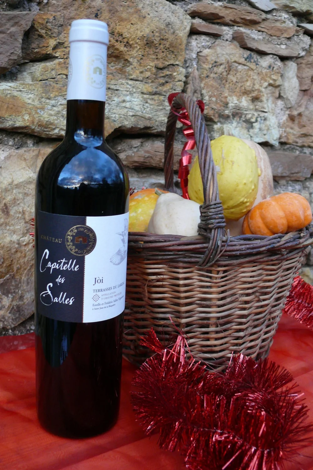 Packshot lifestyle de la cuvee "Joi" (AOP Terrasses du Larzac) : bouteille debout a gauche, etiquette noire "Chateau Capitelle des Salles - Joi - Terrasses du Larzac" bien lisible, panier en osier rempli de courges colorees a droite, guirlande rouge et nappe rouge (ambiance festive). Joli plan de mi