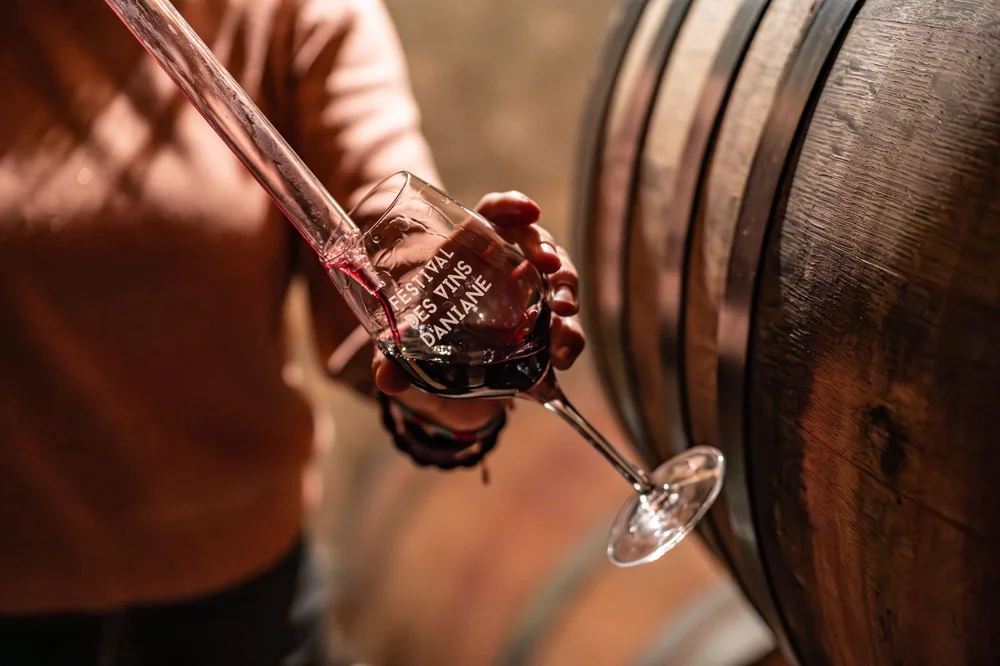 Soutirage du vin rouge à la pipette depuis une barrique, versé dans un verre gravé 'Festival des Vins d'Aniane'. Ambiance chai chaleureuse, gros plan élégant sur les mains et la barrique. Très belle photo de cave. Source : page d'accueil.