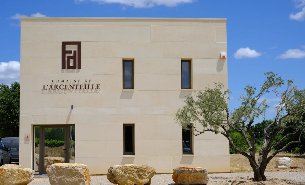Facade du domaine de jour : batiment contemporain en pierre claire sur deux niveaux portant le monogramme et l'enseigne 'Domaine de l'Argenteille', gros rochers et olivier devant, ciel bleu.