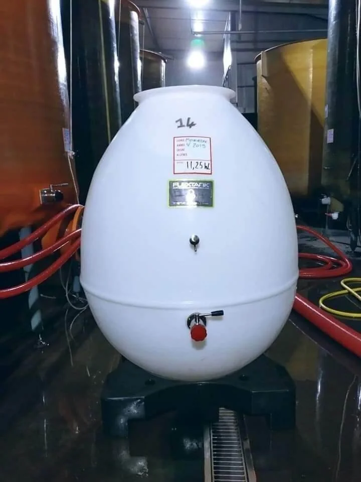 Cuve ovoide blanche (Flextank, 11,25 hl, cuvee 2019) au milieu des cuves du chai, tuyaux rouges au sol. Detail vinification moderne dans la cave.