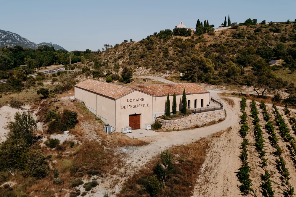 Vue aerienne (drone) du chai du Domaine de l'Eglisette : long batiment ocre avec l'inscription peinte 'DOMAINE DE L'EGLISETTE' sur le mur, cypres, mur de pierres seches, chemin blanc serpentant vers les vignes a droite, collines des Cevennes en arriere-plan et une chapelle perchee sur la crete (l'eg