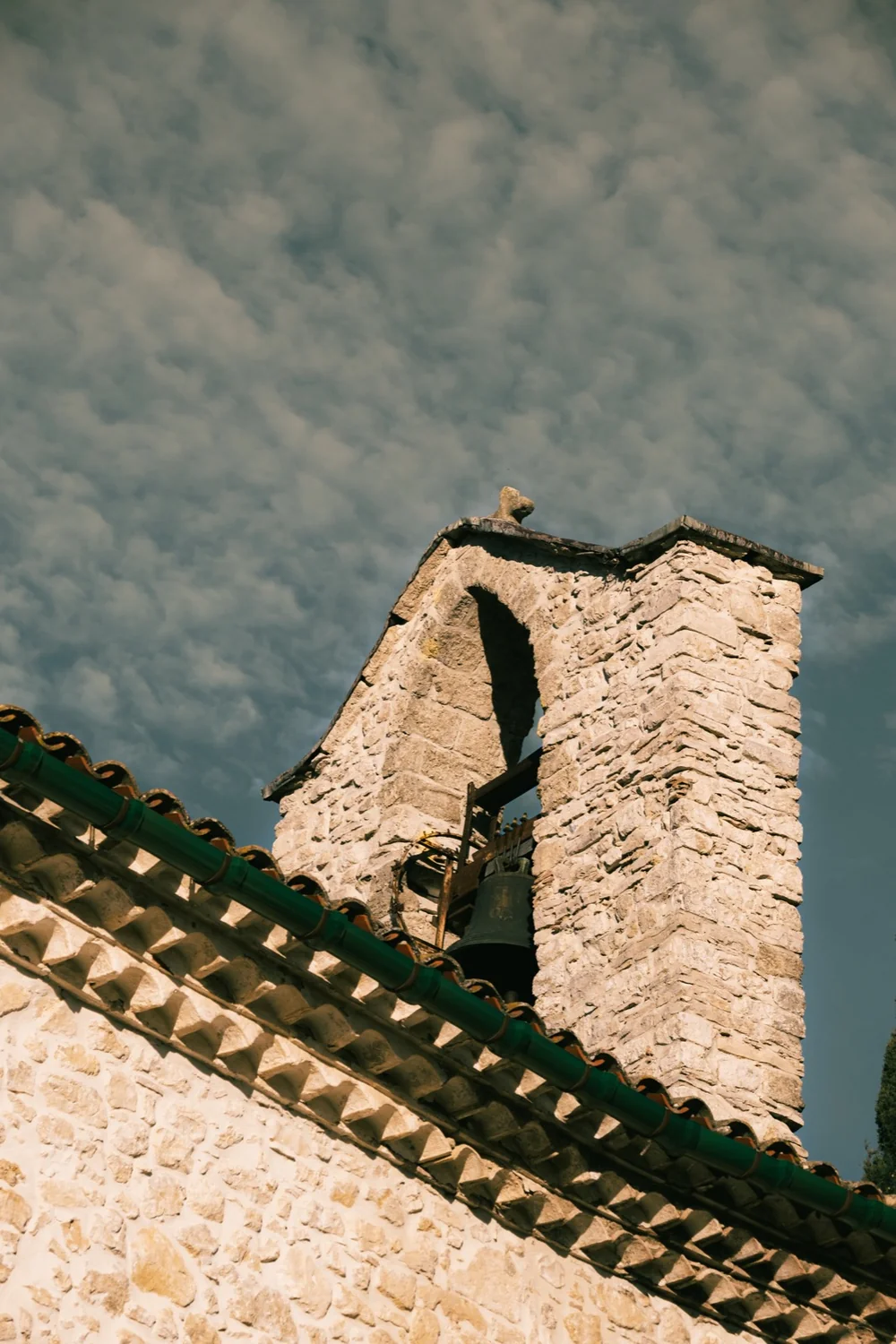 Clocher-mur en pierre sèche du village (La Boissière / Saint-Paul-et-Valmalle) avec sa cloche et son toit de tuiles à faîtière verte, ciel nuageux. Contre-plongée. Ancrage local/patrimonial du domaine, plutôt que bâtiment du domaine lui-même.