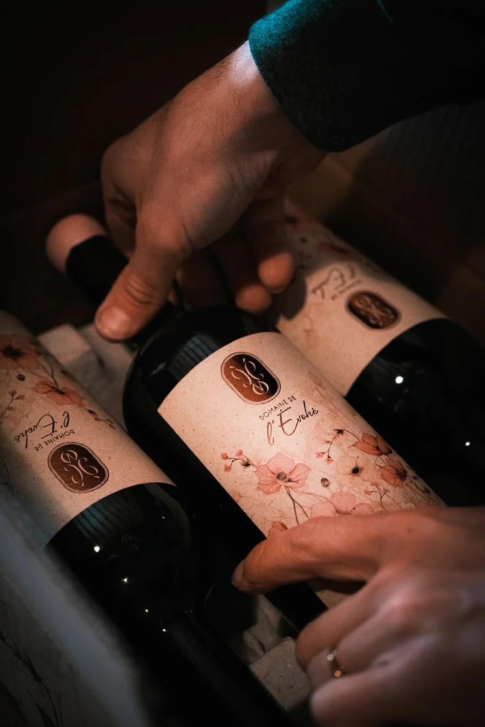 Packshot lifestyle : des mains rangeant trois bouteilles du Domaine de l'Évohé (étiquettes ivoire à motif floral aquarelle, sceau ocre du domaine) dans un carton. Lumière intime et chaleureuse — belle mise en situation, pas une étiquette plate.