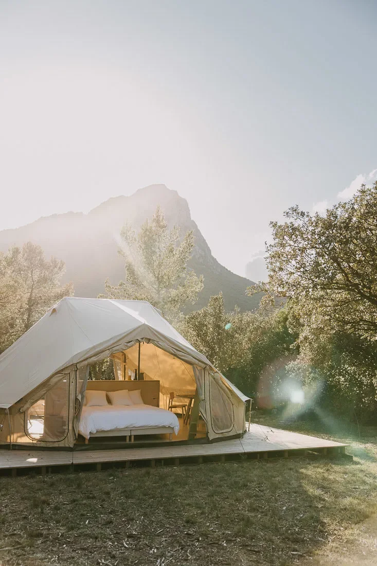 Tente de glamping ouverte sur un lit, baignee de lumiere doree au lever du soleil, Pic Saint-Loup en fond. Image lifestyle de reve pour l'oenotourisme.