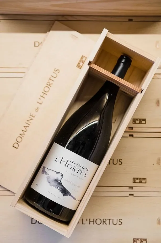 Bouteille Grande Cuvee rouge couchee dans son coffret bois grave 'DOMAINE de l'HORTUS', sur d'autres caisses. Packshot lifestyle haut de gamme (et non etiquette plate).