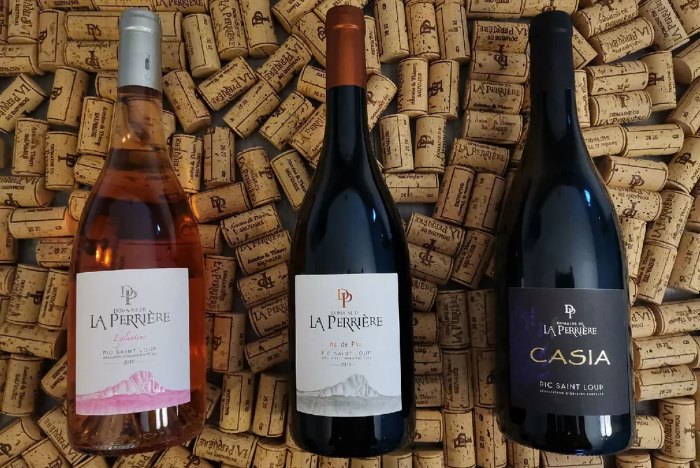 Mise en scene lifestyle : trois bouteilles du domaine (Eglantine rose, As de Pic, Casia rouge - Pic Saint-Loup) posees a plat sur un lit de bouchons de liege graves au nom du domaine. Packshot d'ambiance soigne, pas une simple etiquette.