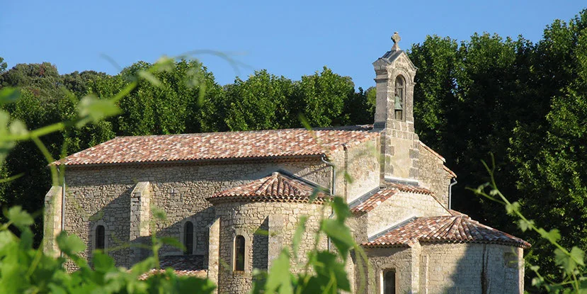 Chapelle/église romane en pierre aux toits de tuiles, clocheton à croix, cadrée à travers les feuilles de vigne sous ciel bleu. Patrimoine bâti du domaine, image patrimoniale et photogénique.