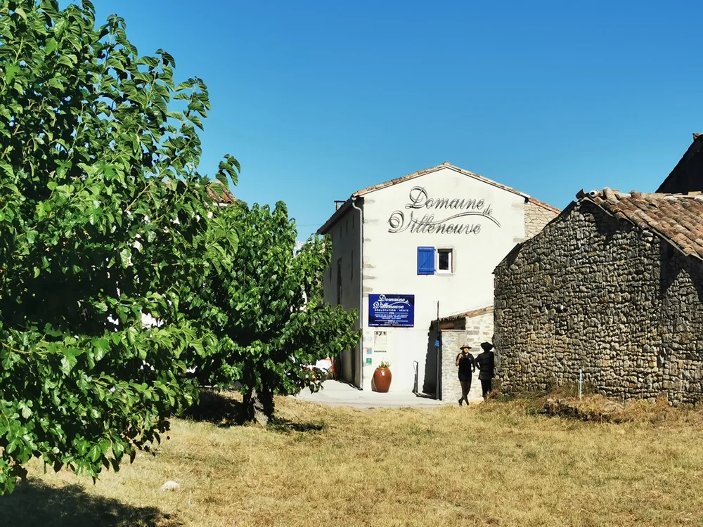 Facade du domaine : batisse en pierre avec 'Domaine de Villeneuve' peint sur le pignon, volets bleus, panneau d'enseigne, jarre et figuier ; deux visiteurs s'approchent de l'entree. Identite des lieux.