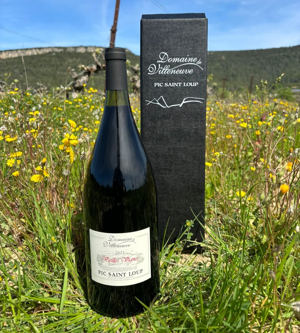 Magnum 'Vieilles Vignes Pic Saint Loup' avec son coffret noir marque 'Domaine de Villeneuve', dans une vigne fleurie de jaune, collines en fond. Lifestyle + packaging premium.