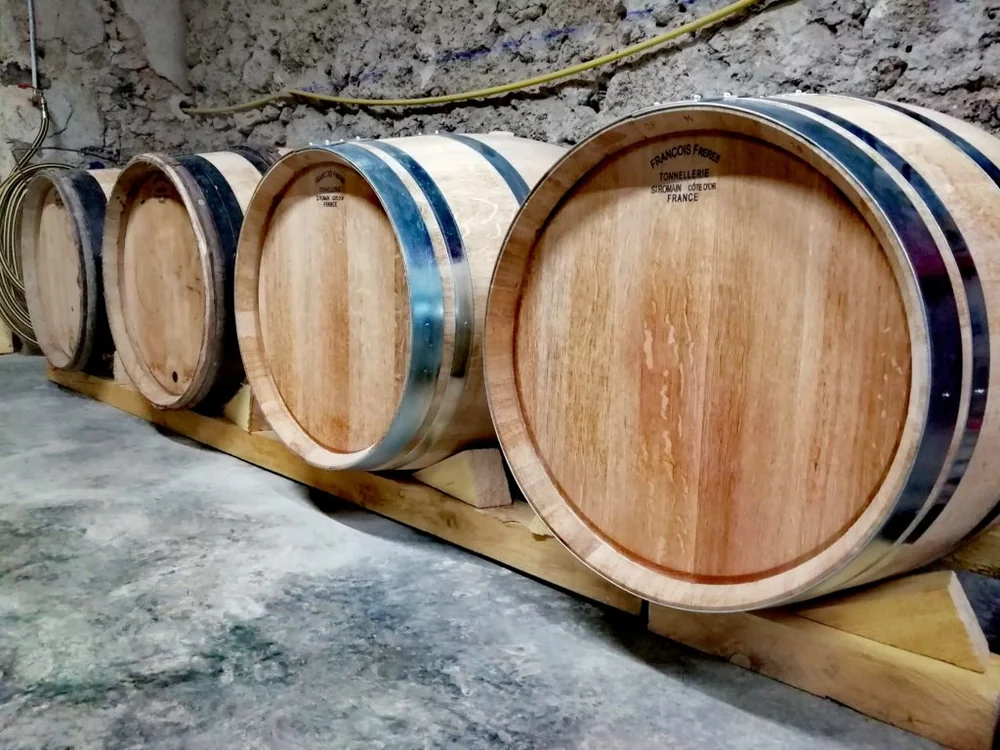 Quatre barriques de chêne (tonnellerie François Frères, Côte-d'Or) alignées sur des supports en bois dans le chai d'élevage aux murs de pierre brute. Authentique, met en valeur l'élevage en fûts (cuvée Cassygre). Belle photo de cave.