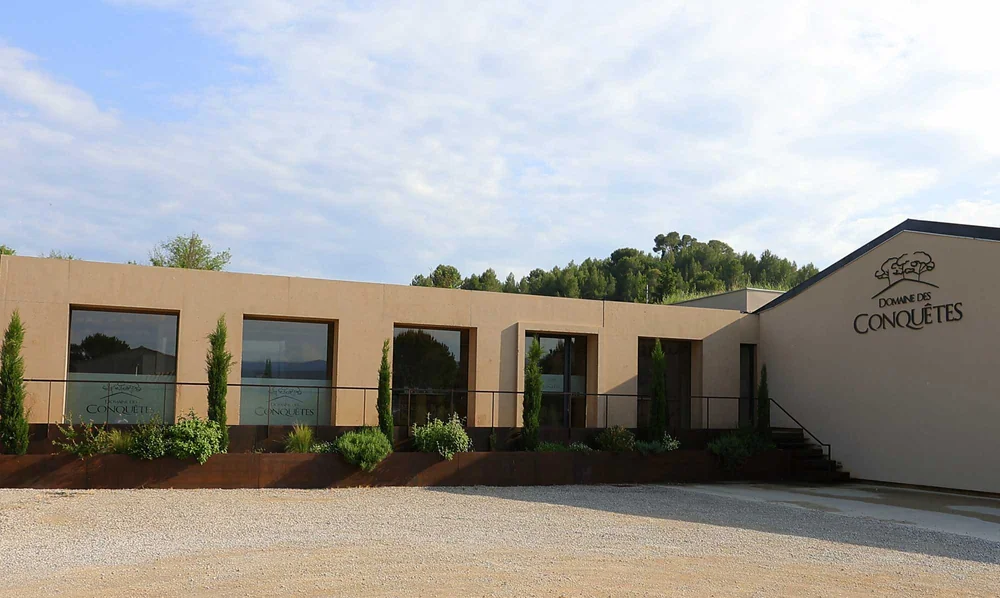 Façade contemporaine du caveau/chai du domaine (inauguré 2019), enduit ocre, grandes baies vitrées, cyprès, et le logo 'Domaine des Conquêtes' gravé sur le pignon. Vue d'ensemble du bâtiment.