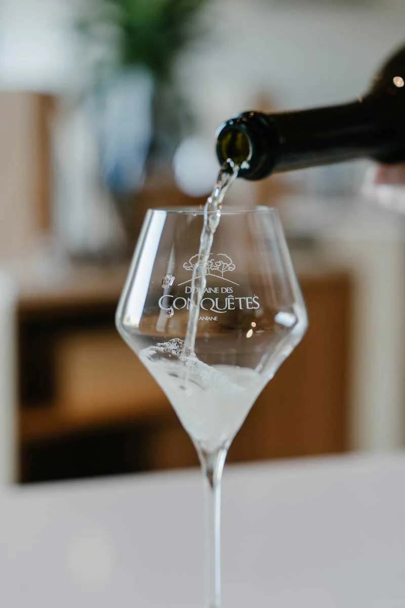 Service du vin : un vin clair versé d'une bouteille dans un verre gravé au logo 'Domaine des Conquêtes Aniane', arrière-plan flou de caveau. Packshot lifestyle élégant et branché.