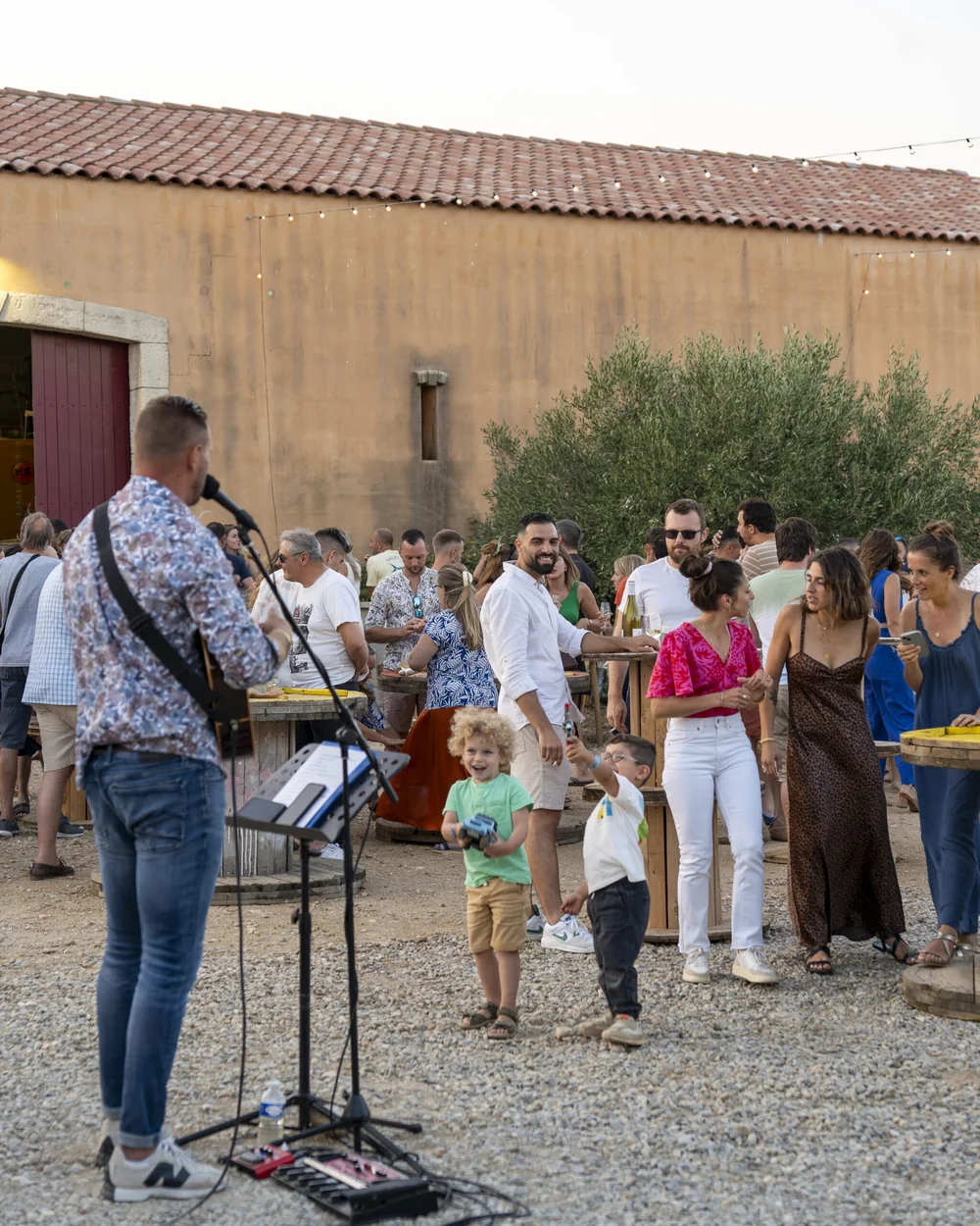 Un musicien joue de la guitare devant le mas ocre (toit en tuiles, porte rouge du chai), foule conviviale avec familles et enfants. Montre le lieu et l'atmosphere chaleureuse.
