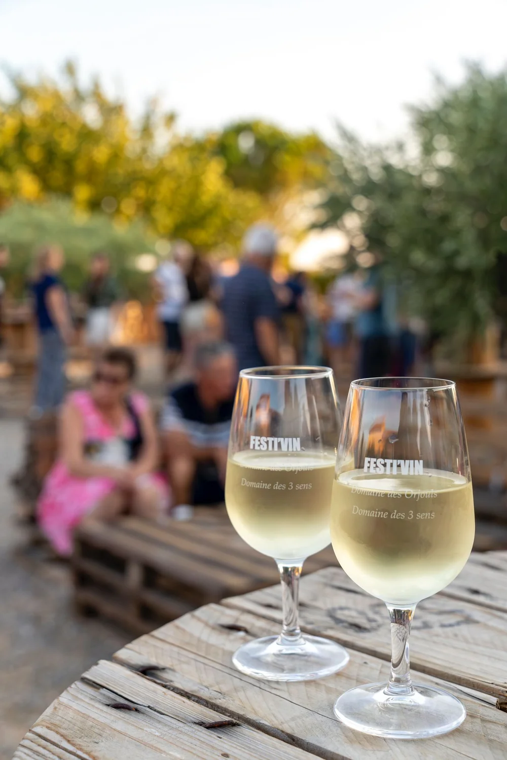 Deux verres de vin blanc graves 'FESTI'VIN / Domaine des Orjouls' sur une table en touret, bokeh heure doree avec des gens en fond. Tres atmospherique.