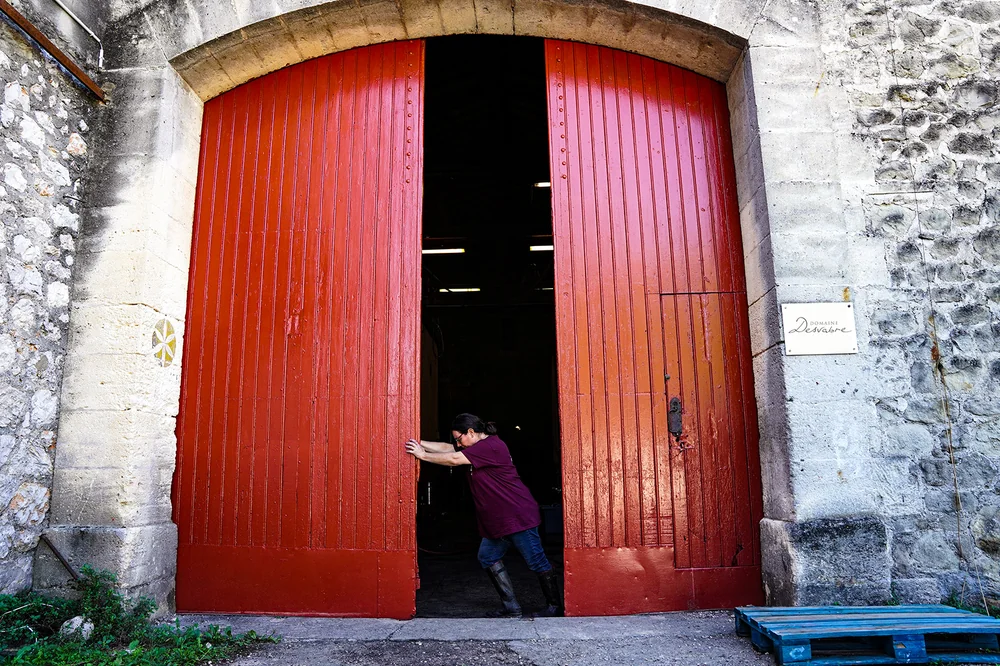 Entrée du chai/cave : grande double porte en bois rouge vif encadrée de pierres de taille anciennes, une personne en train d'ouvrir le battant. Élément architectural identitaire du mas. Cadrage un peu serré mais caractéristique du domaine. 1620x1080.