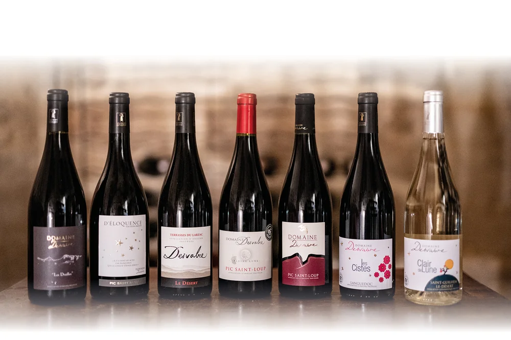 Photo lifestyle de la gamme complète : sept bouteilles Domaine Desvabre alignées (Les Diavlès, D'Éloquence, Le Désert Terrasses du Larzac, Pleine Lune Pic Saint-Loup, un Pic Saint-Loup, Les Cistes Languedoc, et le blanc Clair de Lune Saint-Guilhem-le-Désert) sur un comptoir bois, fond de chai flou.
