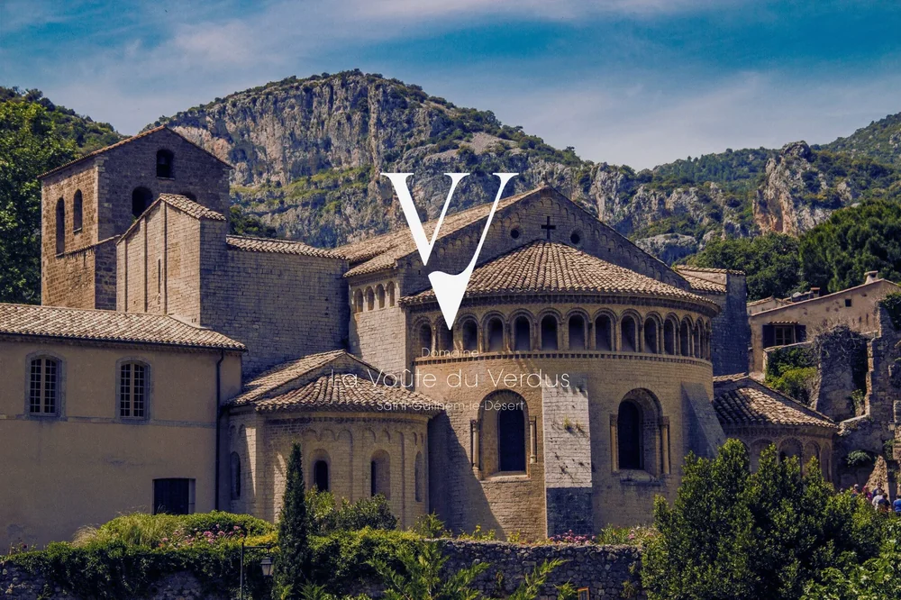 L'Abbaye de Gellone de Saint-Guilhem-le-Desert (site UNESCO) dominee par les falaises, cadre emblematique du domaine. Discret filigrane du logo VV en surimpression. Beau visuel d'identite de lieu, format paysage.