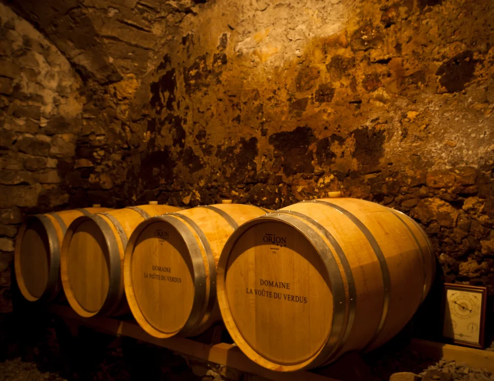 Rangee de barriques marquees 'DOMAINE LA VOUTE DU VERDUS' dans une cave voutee en pierre, eclairage chaud dore. Superbe image du chai a barriques, identitaire et atmospherique.