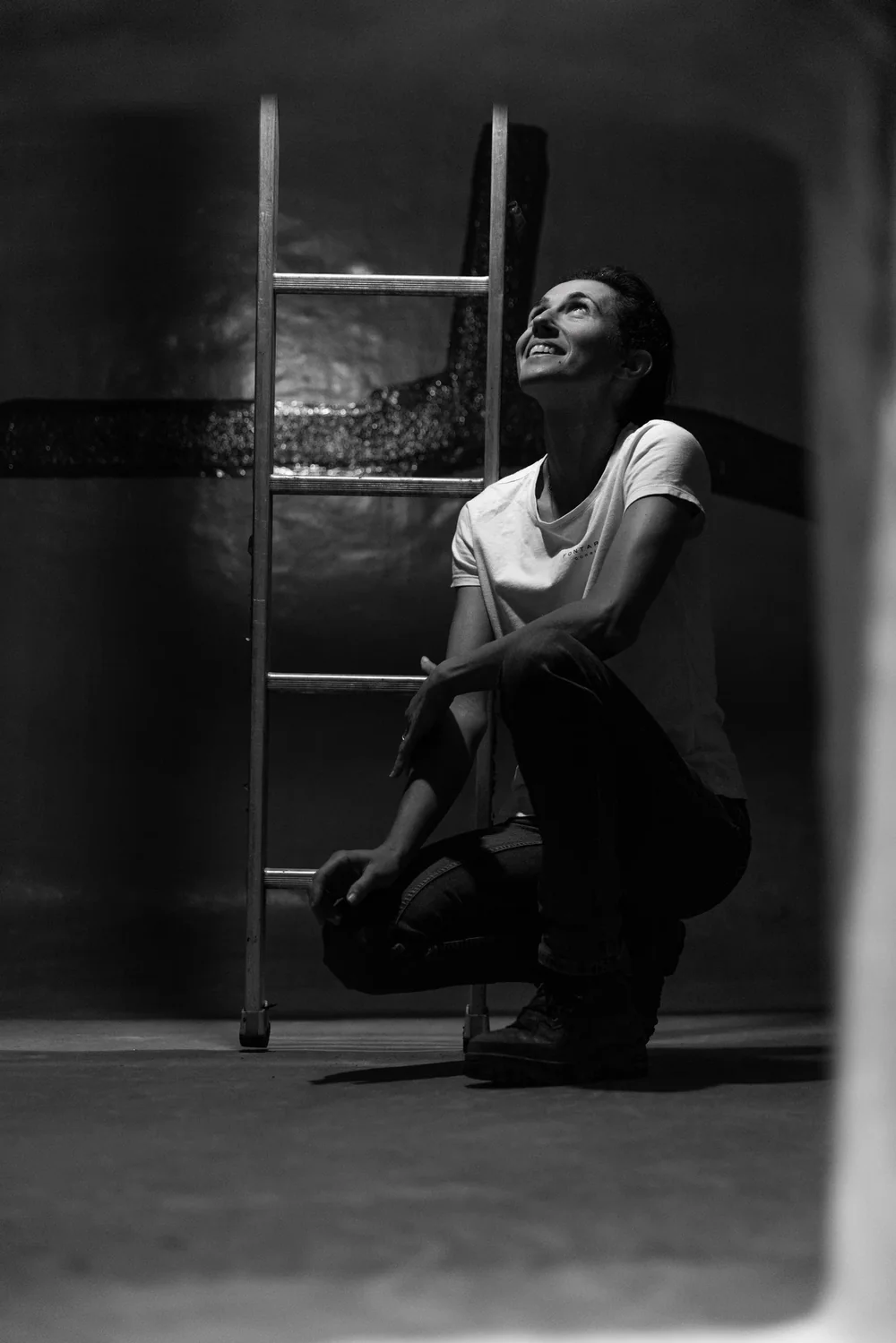 Portrait noir et blanc artistique de Melanie accroupie pres d'une echelle, contre une cuve, le regard leve. Tres beau portrait d'ambiance qui humanise le domaine.