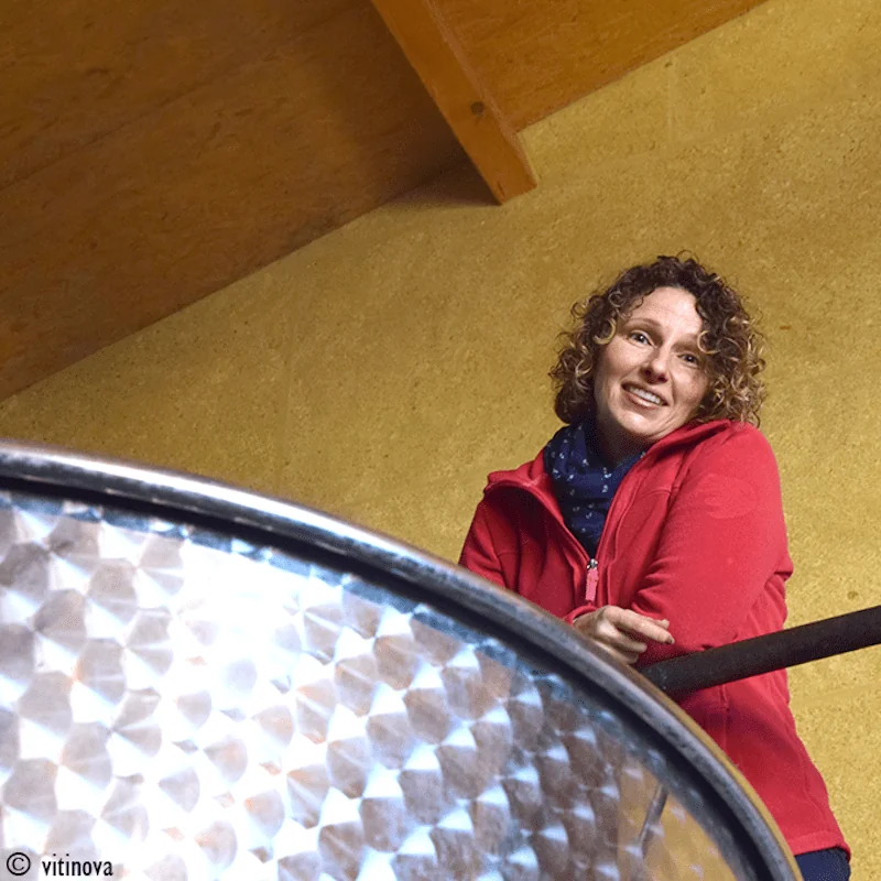 Portrait d'Audrey Bonnet-Koenig (directrice generale du domaine), souriante, vetue d'une veste rouge et d'une echarpe, penchee au-dessus d'une grande cuve en inox dans le chai. Vue plongeante authentique qui humanise la marque et associe la vigneronne au travail de vinification. Image phare du HUMAI