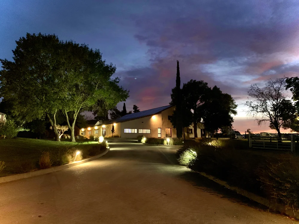 Le mas a la tombee de la nuit : allee d'acces eclairee par bornes lumineuses, batiment au toit en pente, cypres et arbres se decoupant sur un ciel crepusculaire rose et bleu. Image tres atmospherique et valorisante du domaine.