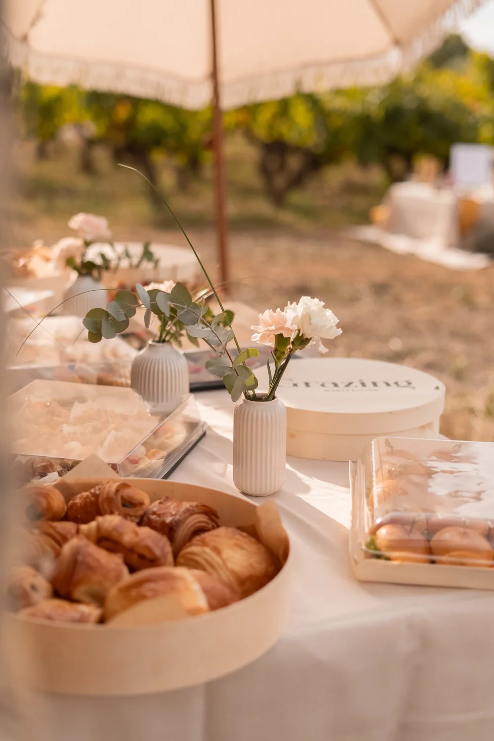 Table de reception/brunch dressee en exterieur dans les vignes : viennoiseries, vases de fleurs, parasol a franges, lumiere doree. Tres atmospherique, valorise l'offre evenementielle dans le cadre du vignoble.