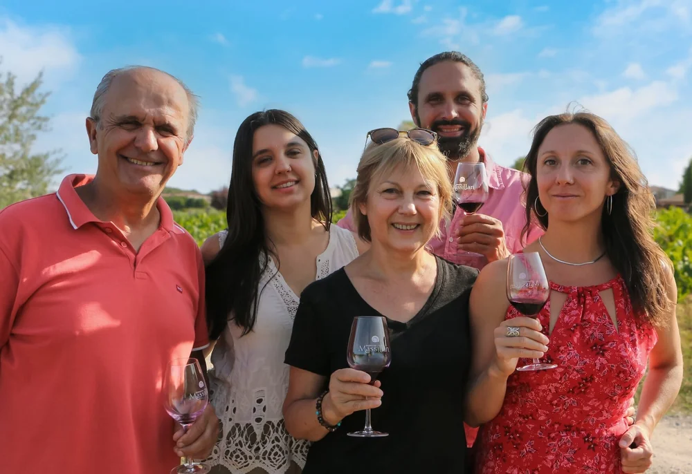 Portrait de la famille Reboul (vignerons actuels, 7e generation) dans les vignes au soleil, chacun tenant un verre de vin grave au logo Massillan. Cinq personnes souriantes, ambiance chaleureuse et authentique. Image phare pour incarner l'humain et la transmission familiale.