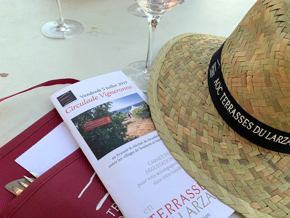 Scene lifestyle de degustation/evenement : chapeau 'AOC Terrasses du Larzac', carnet 'Circulade Vigneronne 2019' et verres de vin sur une table. Evoque les evenements et l'appellation.