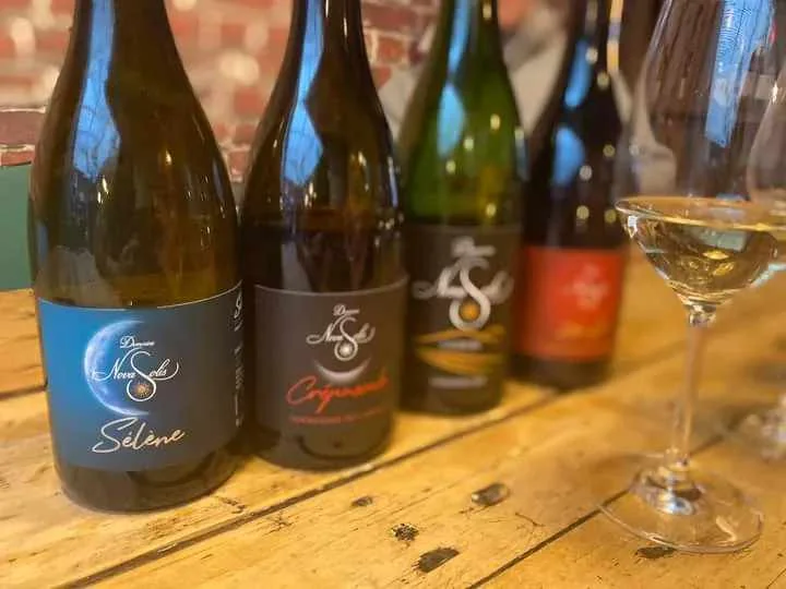 Alignement des bouteilles du domaine (Selene, Crepuscule...) en situation sur une table en bois, mur de briques en fond, un verre de vin blanc a droite. Packshot lifestyle, ambiance chaleureuse (pas une etiquette plate).