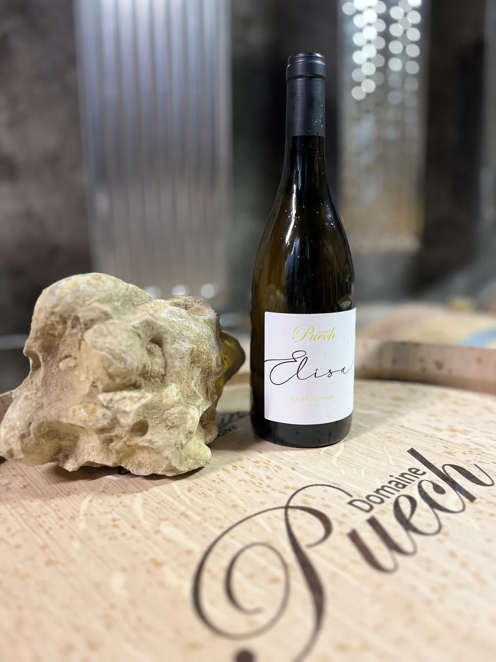 Bouteille de Chardonnay 'Elisa' posee sur une tete de barrique gravee 'Domaine Puech', pierre calcaire de terroir a cote, cuves inox en arriere-plan. Met en valeur le chai et le savoir-faire.