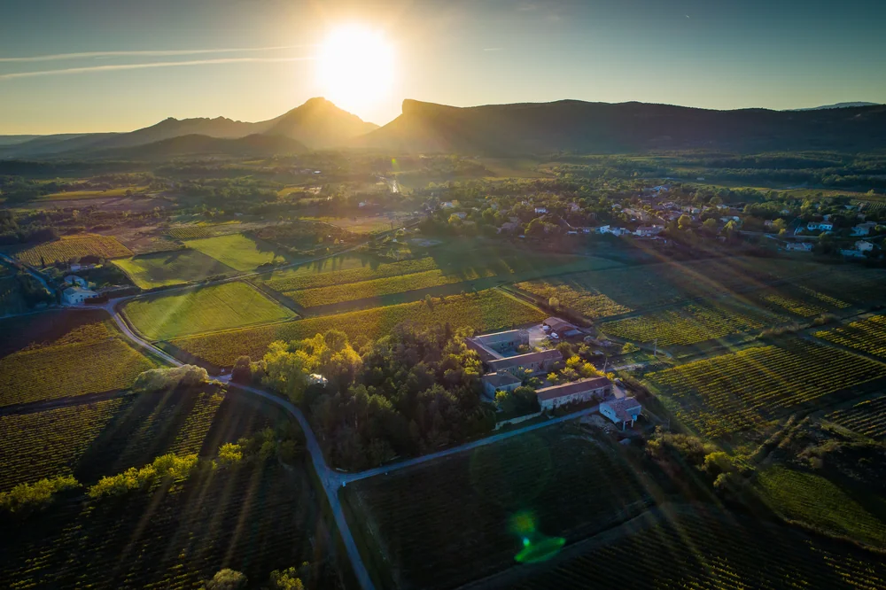 Magnifique vue aerienne au coucher du soleil sur le terroir du Domaine UMA a Valflaunes : au premier plan le mas/bati du domaine niche dans les arbres, entoure de parcelles de vignes en mosaique, et a l'arriere-plan la silhouette emblematique du Pic Saint-Loup et de l'Hortus baignee dans la lumiere