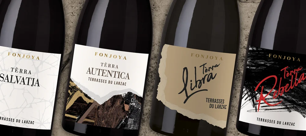 Composition lifestyle des quatre cuvees de la gamme Terra (Terrasses du Larzac : Salvatja, Autentica, Libra, Rebella) alignees sur une surface de pierre, gros plan sur les etiquettes elegantes. Belle mise en situation des bouteilles haut de gamme.