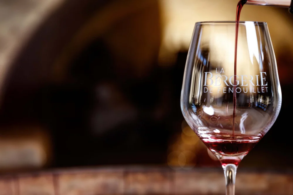 Photo héros premium : vin rouge versé dans un verre gravé au logo « La Bergerie de Fenouillet », lumière chaude, arrière-plan flou de barriques dans le caveau voûté. Composition et qualité professionnelles (2881x1920).
