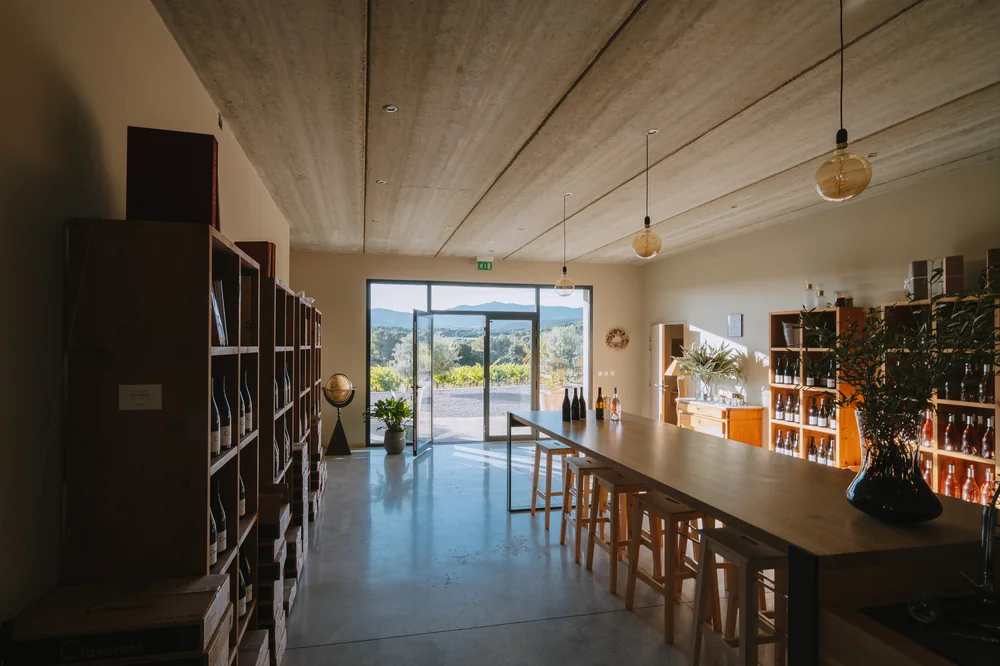 Le caveau de degustation: longue table en bois clair et tabourets, plafond bois, suspensions design, etageres de bouteilles, grande baie vitree ouvrant sur les collines. Espace d'accueil chaleureux et haut de gamme. ALT: 'Caveau de degustation La Closeraie accueil degustation groupes'.