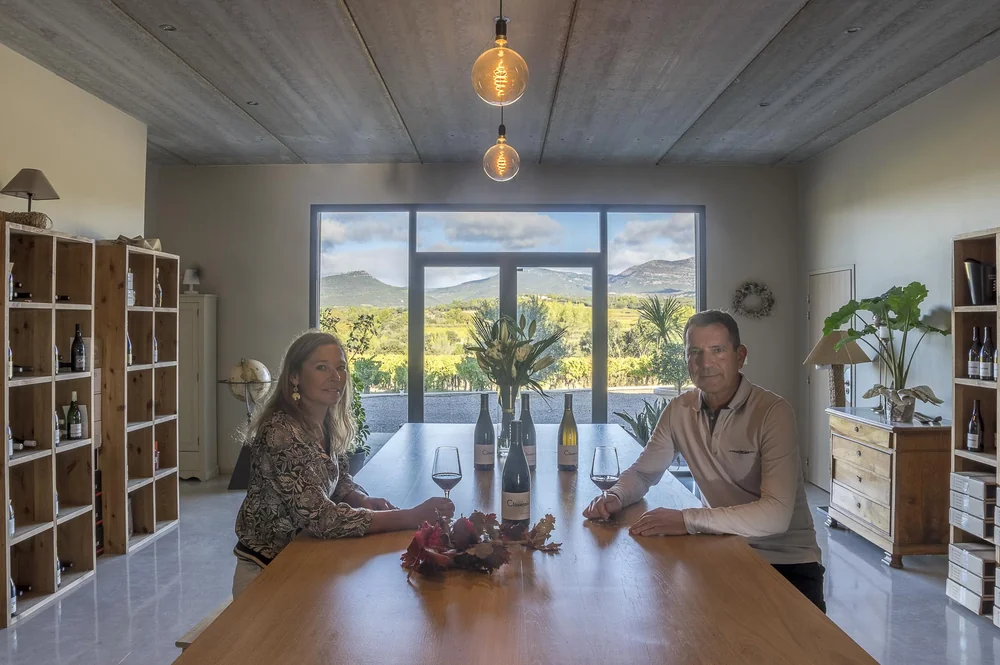 Paul et Marie Ryckwaert, les vignerons, assis face a face a la grande table du caveau de degustation. Bouteilles et verres de vin sur la table, grande baie vitree ouvrant sur les vignes et les collines du Larzac, suspensions design. Portrait lifestyle chaleureux et premium, incarne parfaitement le c