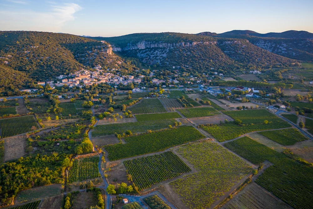 Vue aérienne au couchant du village de Corconne blotti sous les falaises calcaires, entoure d'un patchwork de parcelles de vignes. Photo drone haute resolution (4000x2665), lumiere doree, composition spectaculaire : l'image signature du terroir.