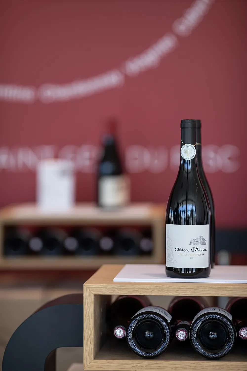 Mise en situation lifestyle : bouteille de Chateau d'Assas posee sur un casier a vin en bois dans le caveau, mur bordeaux en fond. Packshot d'ambiance soigne (pas une etiquette plate).